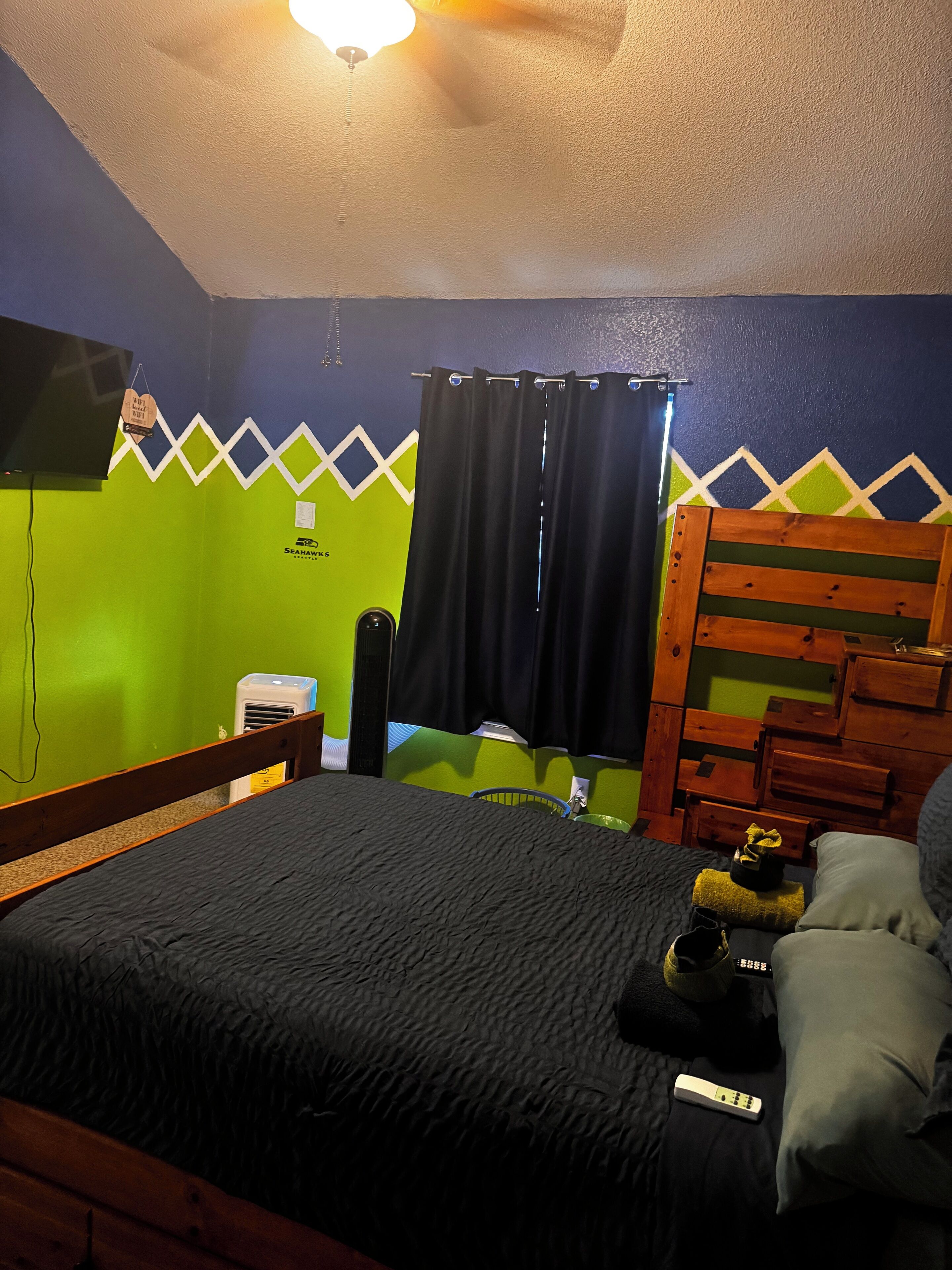 Un dormitorio con cama, cabecera de madera, ventilador de techo, televisión montada en la pared, calefactor y ventana con cortinas.