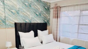 Una cama bien hecha con sábanas blancas y un detalle azul, una mesita de noche con una lámpara y una pared con estampado de mármol.