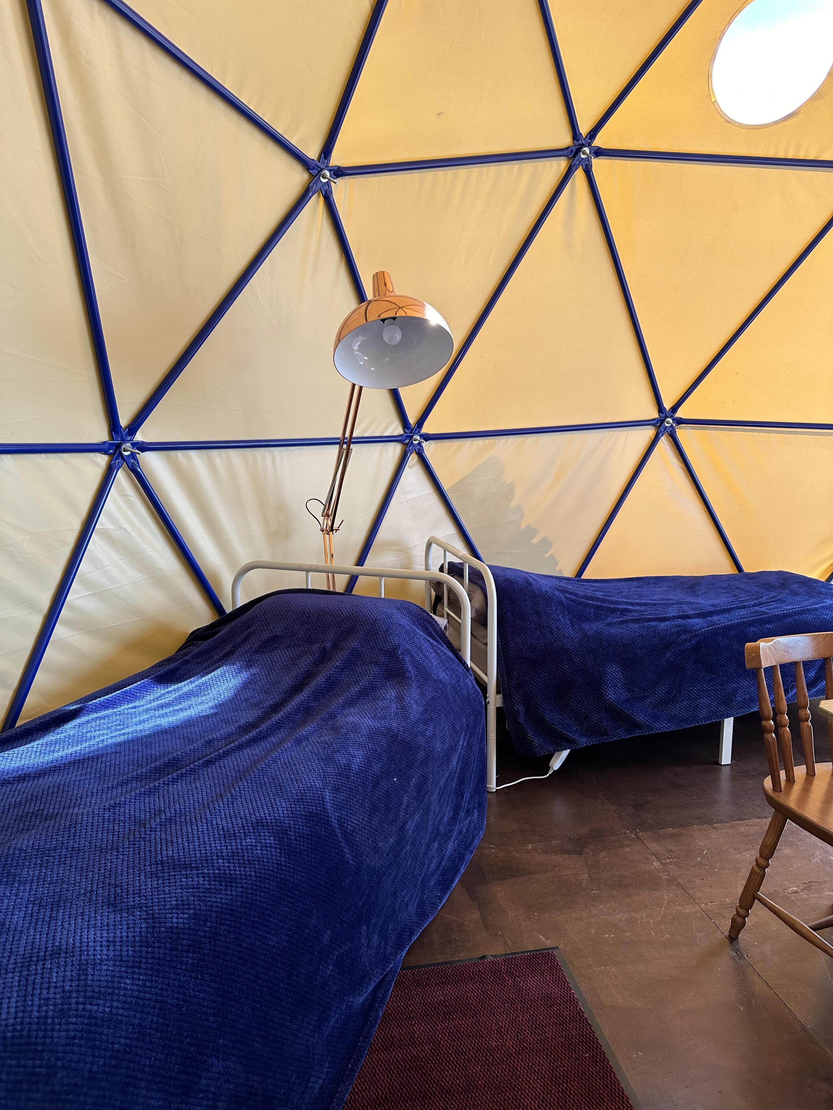 Una tienda de campaña con un estampado geométrico, dos camas con ropa de cama azul, una lámpara colgante y una silla de madera.