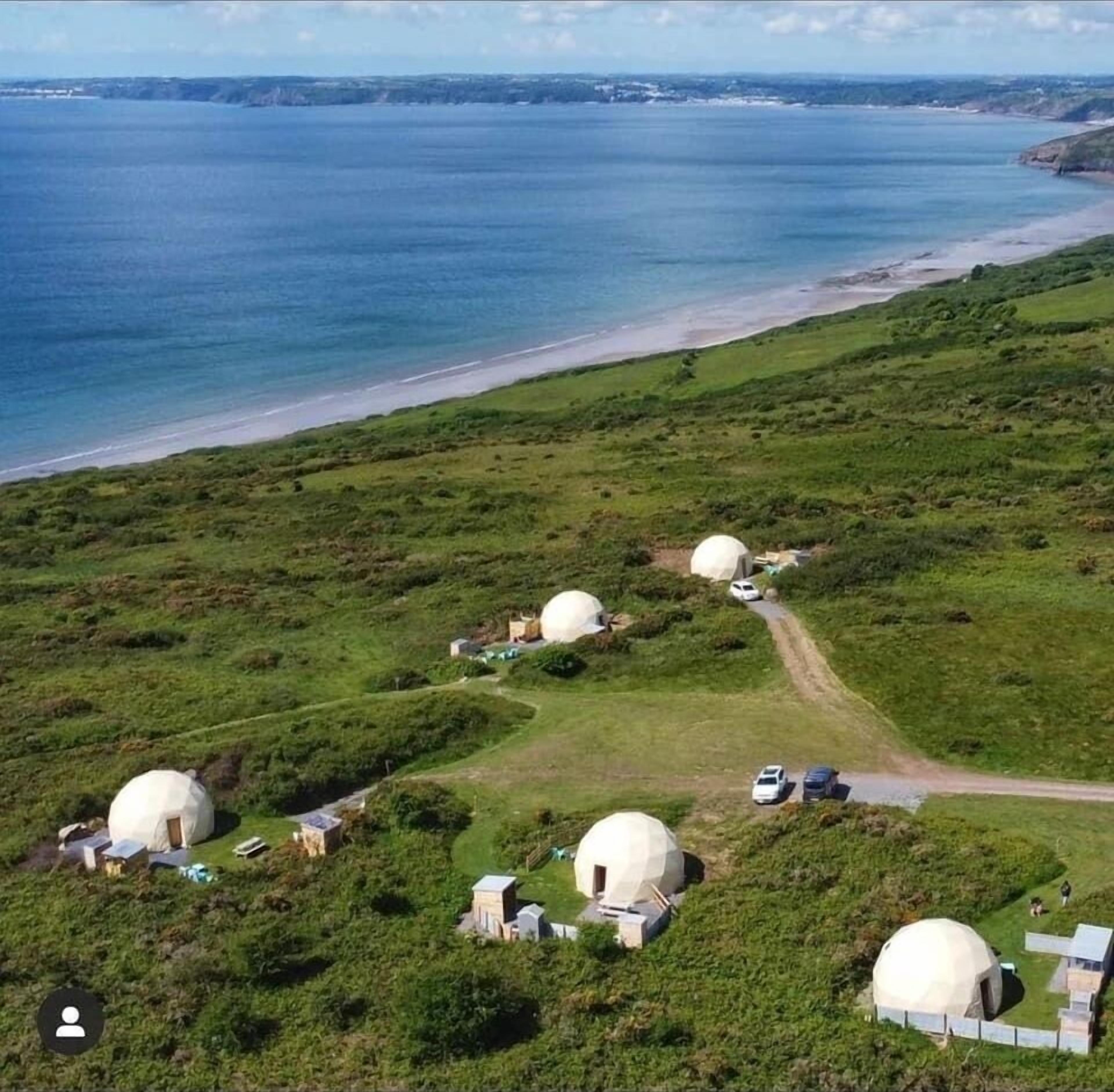 Een kustgebied met meerdere koepelvormige gebouwen, waarschijnlijk verblijfplaatsen, omringd door groene velden en een strand.