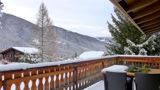 En balkon med trægelænder, et bord og stole, med udsigt over et snøklædt bjerglandskab.