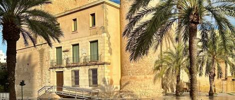 A historic building with a stone facade, a balcony, and palm trees in the foreground.