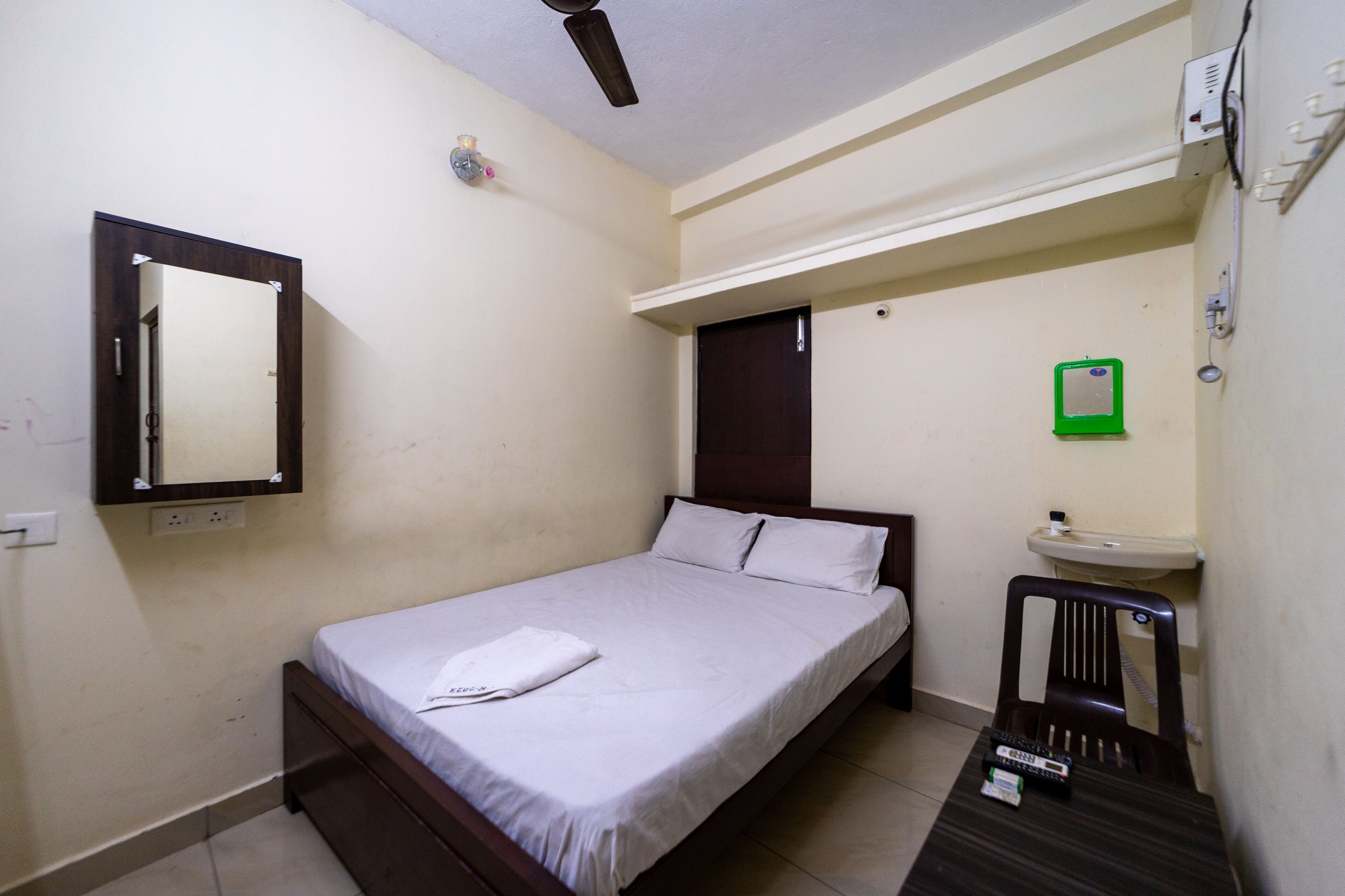 A single bed with a white mattress and pillows, a small bedside table with a remote control, a wall-mounted mirror, a ceiling fan, a green emergency exit sign, and a small sink.