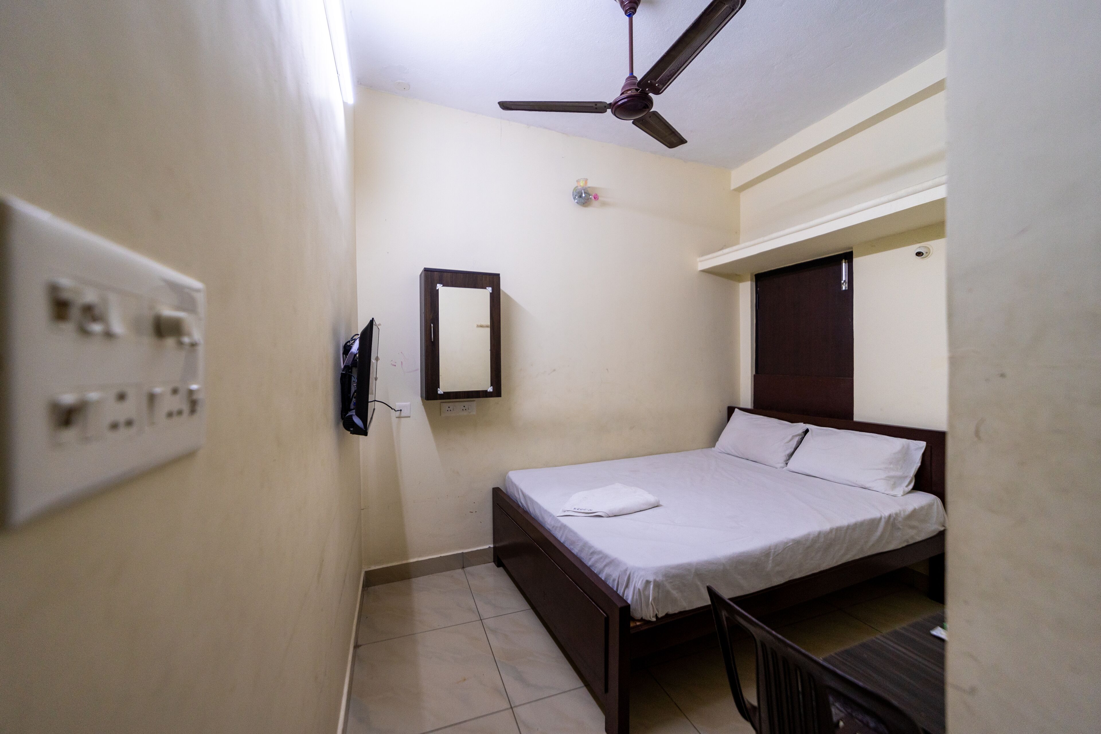 A small, single-bed room with a ceiling fan, a wall-mounted TV, and a power outlet.