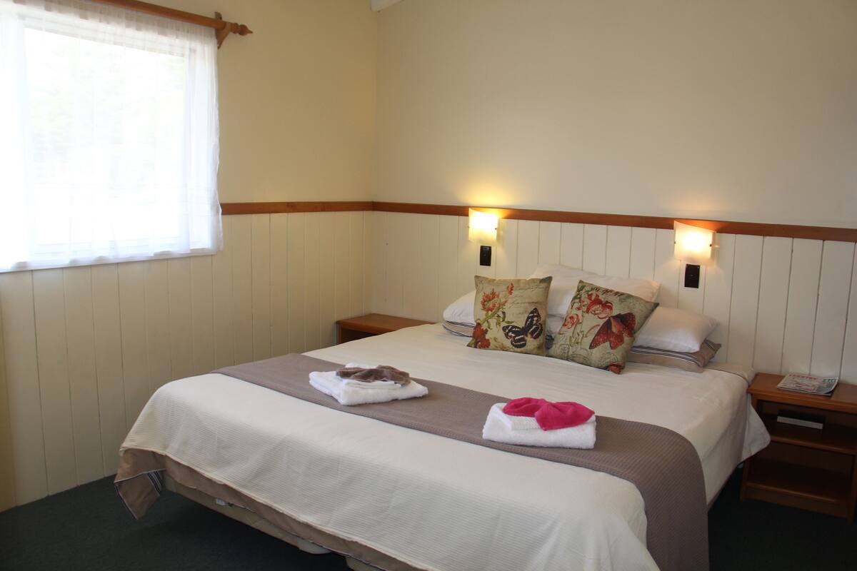 A neatly made bed with a brown headboard, two bedside lamps, and a window with sheer curtains.