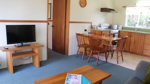 Un espacio habitable compacto con una televisión sobre un soporte de madera, una mesa de comedor con sillas, un sofá y una zona de cocina con estufa y fregadero.