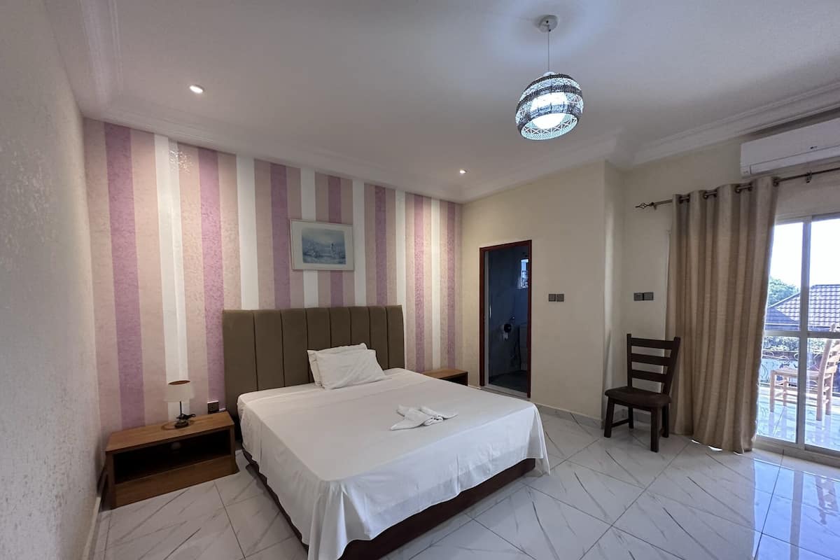 Habitación de hotel con una cama grande, una mesita de noche con lámpara, una silla, una puerta y vistas al exterior.