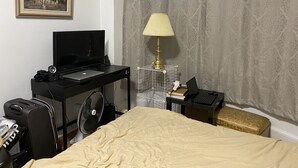 Una habitación de hotel con una cama, un escritorio con una computadora portátil, un televisor, una lámpara, una maleta y un cuadro en la pared.
