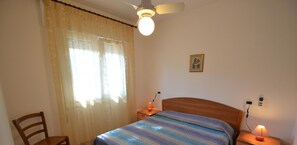 Een slaapkamer met een bed, een stoel, een nachtkastje met een lamp, een plafondventilator en een raam met gordijnen.