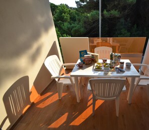 Een balkon met een tafel voor vier personen, omringd door witte stoelen. Op de tafel staan diverse items, waaronder een doos met de tekst 'Alparco', een theepot, kopjes en een fles.