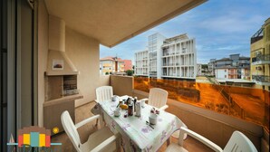 A balcony with a table set for two, chairs, a grill, and a view of surrounding buildings. - Apartment with 2 bedrooms near to the beach LP 10 (Rosolina Mare)