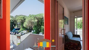 Varanda com vista para carros estacionados e árvores, um sofá e uma porta que dá para um quarto.