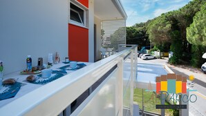 Um edifício moderno com varanda, área de refeições com itens de café da manhã e vista para a rua com carros estacionados e árvores.