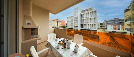 Un balcón con una mesa para dos, sillas, una parrilla y vistas de edificios circundantes.