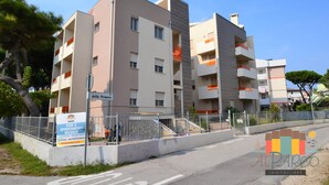 Een meerlaags woongebouw met balkons, een bord met een advertentie voor vakantieappartementen, en het logo van ALPARCO IMMOBILIARE.