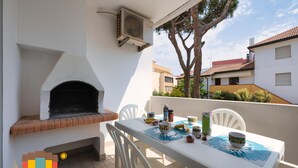 Une terrasse extérieure avec une table blanche dressée pour le repas, une cheminée en briques et des chaises blanches.