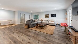 Een ruime woonkamer met een grijze bank, een witte fauteuil en een houten salontafel. Er staat een rode poef en een klein tafeltje met een lamp. De kamer heeft houten vloeren en lichtblauwe wanden.