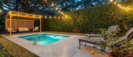 Un jardin avec une piscine, un espace salon couvert et des guirlandes lumineuses.