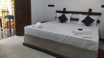 Una cama bien hecha con sábanas blancas y dos almohadones decorativos, un cabecero de madera y una pequeña mesa con una silla al fondo.