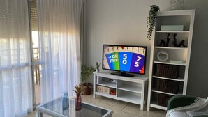 Ein Wohnzimmer mit Fernseher, einem Glastisch, einem grünen Sofa, einer weißen Regaleinheit und Blick durch ein Fenster mit leichtem Vorhang.