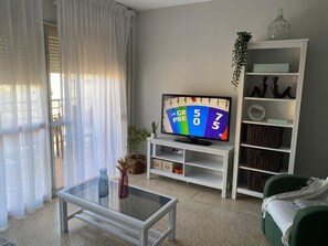 Een woonkamer met een televisie, een glazen salontafel, een groene bank, een witte boekenkast en uitzicht door een raam met dunne gordijnen.