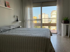 Une chambre avec un grand lit, une vue sur les immeubles par la fenêtre et un balcon.