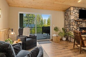 A cozy living area with a fireplace, a stone wall, and a view of trees through a sliding glass door. - Mill Creek Rd Lakehouse (Ronald)