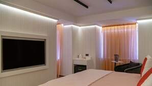 A modern hotel room with a large flat-screen TV mounted on the wall, a bedside table with a lamp, a chair, and a bed with a red and white pillow. - Royal Teos Thermal Resort Clinic & Spa - Halal Hotel (Seferihisar)