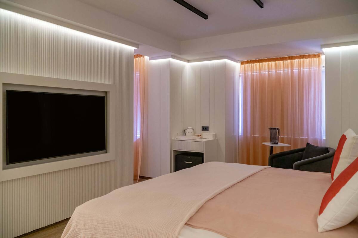 Une chambre d’hôtel moderne dotée d’un grand téléviseur à écran plat fixé au mur, d’une table de chevet avec une lampe, d’une chaise et d’un lit avec un oreiller rouge et blanc.