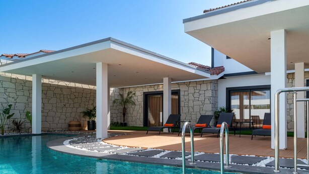 A pool area with a stone wall, lounge chairs, and a covered patio. - Royal Teos Thermal Resort Clinic & Spa - Halal Hotel (Seferihisar)