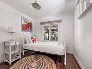 A bedroom with a white bed, a bedside table, a lamp, a colorful rug, and a window with a view of trees. - Wisteria Vines Estate—Rustic Elegance by the Coast (Wilyabrup)