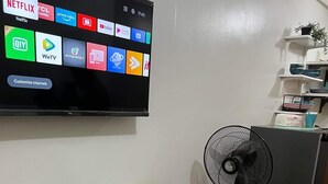 A room with a flat-screen TV displaying various streaming services, a table with a fan, and a refrigerator.
