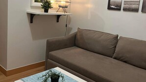 A living room with a brown sofa, a small white table with a plant, a wall-mounted shelf with two lamps, and a mirror above the shelf.