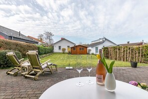 A patio with outdoor furniture, a table with wine glasses and a bottle, and a view of a house in the background.