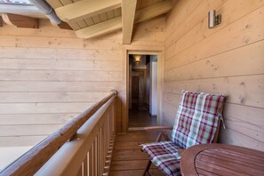 Interior de una cabaña de madera con balcón, un sillón a cuadros y una mesa de madera.