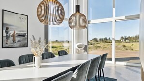 A modern dining area with a large table, chairs, and pendant lights. A framed picture is on the wall, and there is a view of the outdoors through large windows.