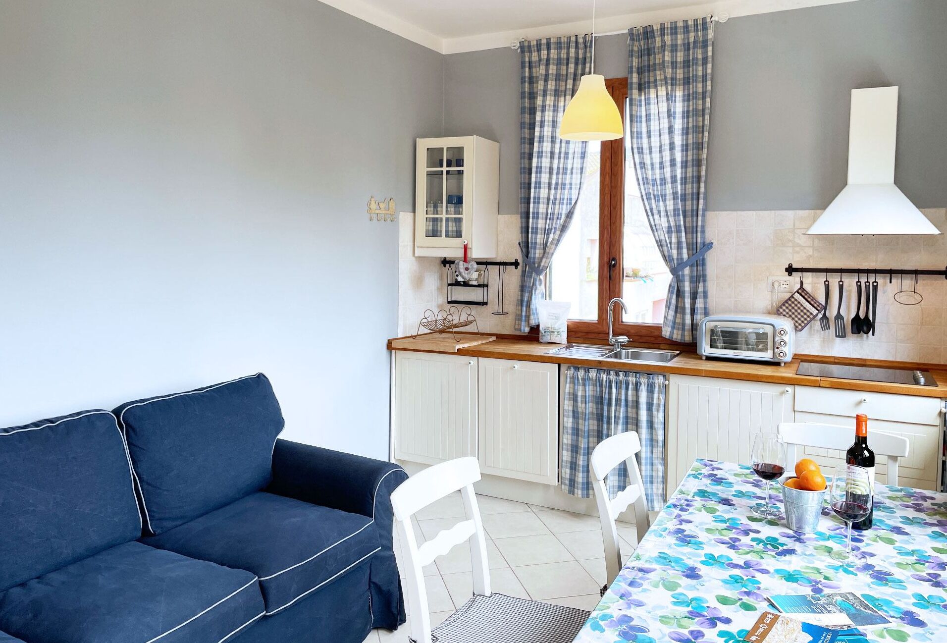 Een kleine keuken met een eethoek, met een bank, stoelen, een tafel met een bloemkleurig tafelkleed, een broodrooster en een hangende lamp.
