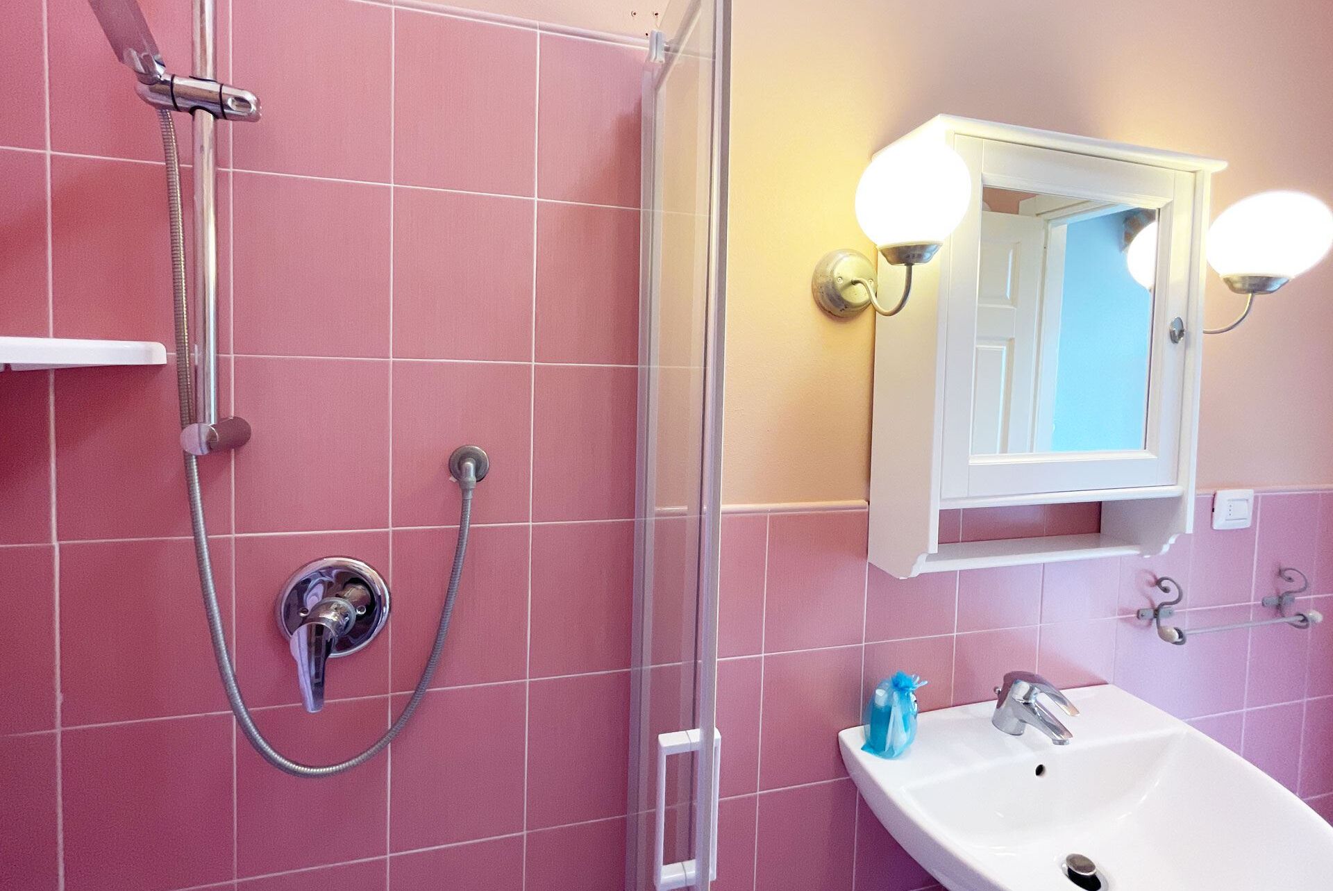 A bathroom with pink tiled walls, a shower with a handheld showerhead, a sink with a mirror above it, and two wall-mounted lights.