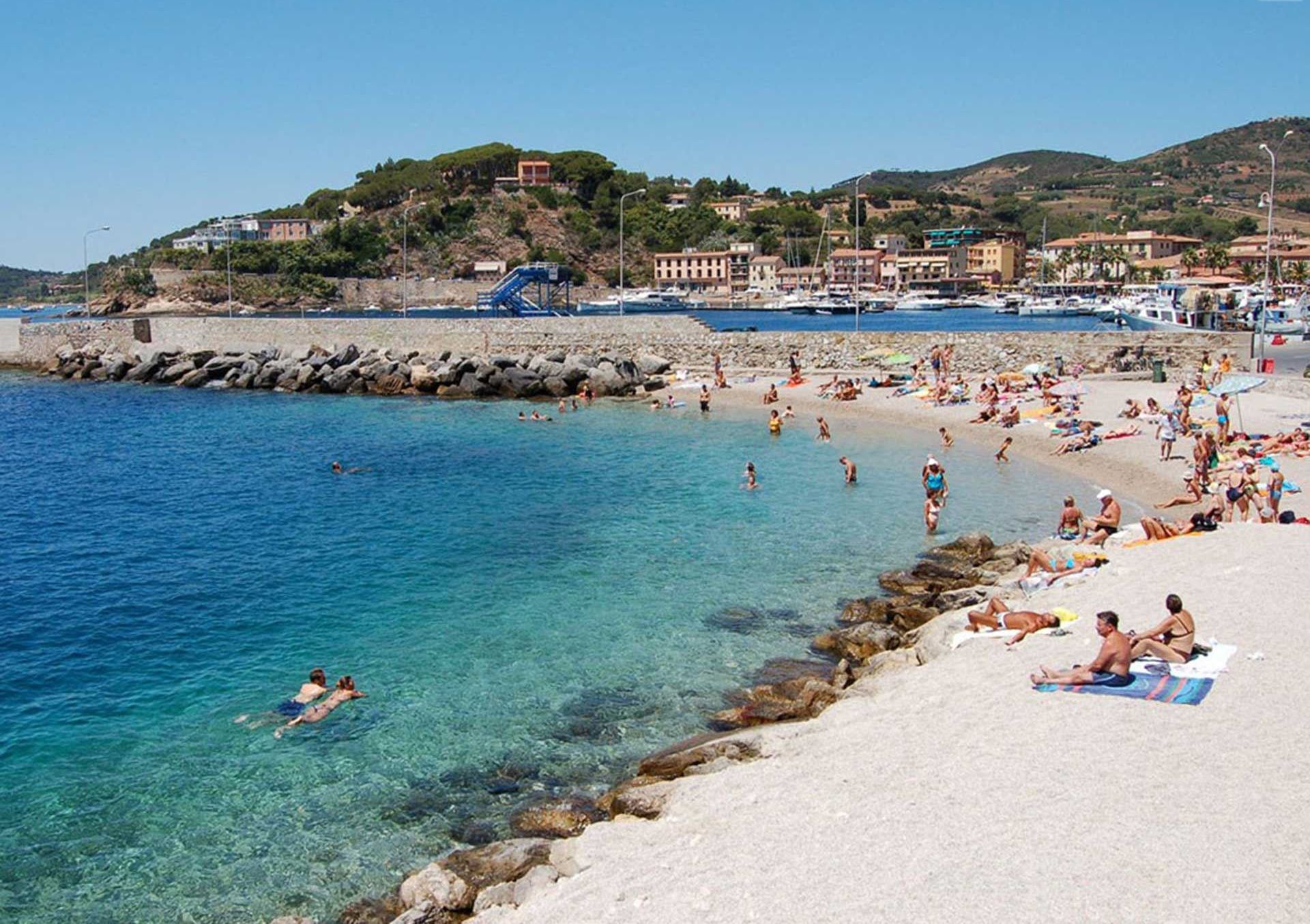 Une plage avec une eau d’un bleu limpide, des gens qui se baignent et prennent des bains de soleil, et des bâtiments en arrière-plan.