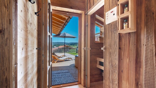 Un interior de madera con un vidrio que da a un patio exterior con terraza de madera, sillones de descanso y un cielo azul despejado.