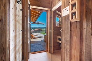 A wooden interior with a glass door leading to an outdoor patio with a wooden deck, lounge chairs, and a clear blue sky.