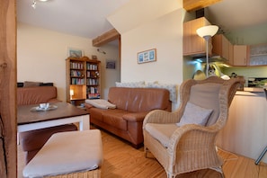 A cozy living room with a brown leather sofa, a wicker armchair, a wooden table, and a bookshelf. - Apartment "Poggenkrug Halbsguth" with Lake View, Private Terrace & Wi-Fi (Wolgast)