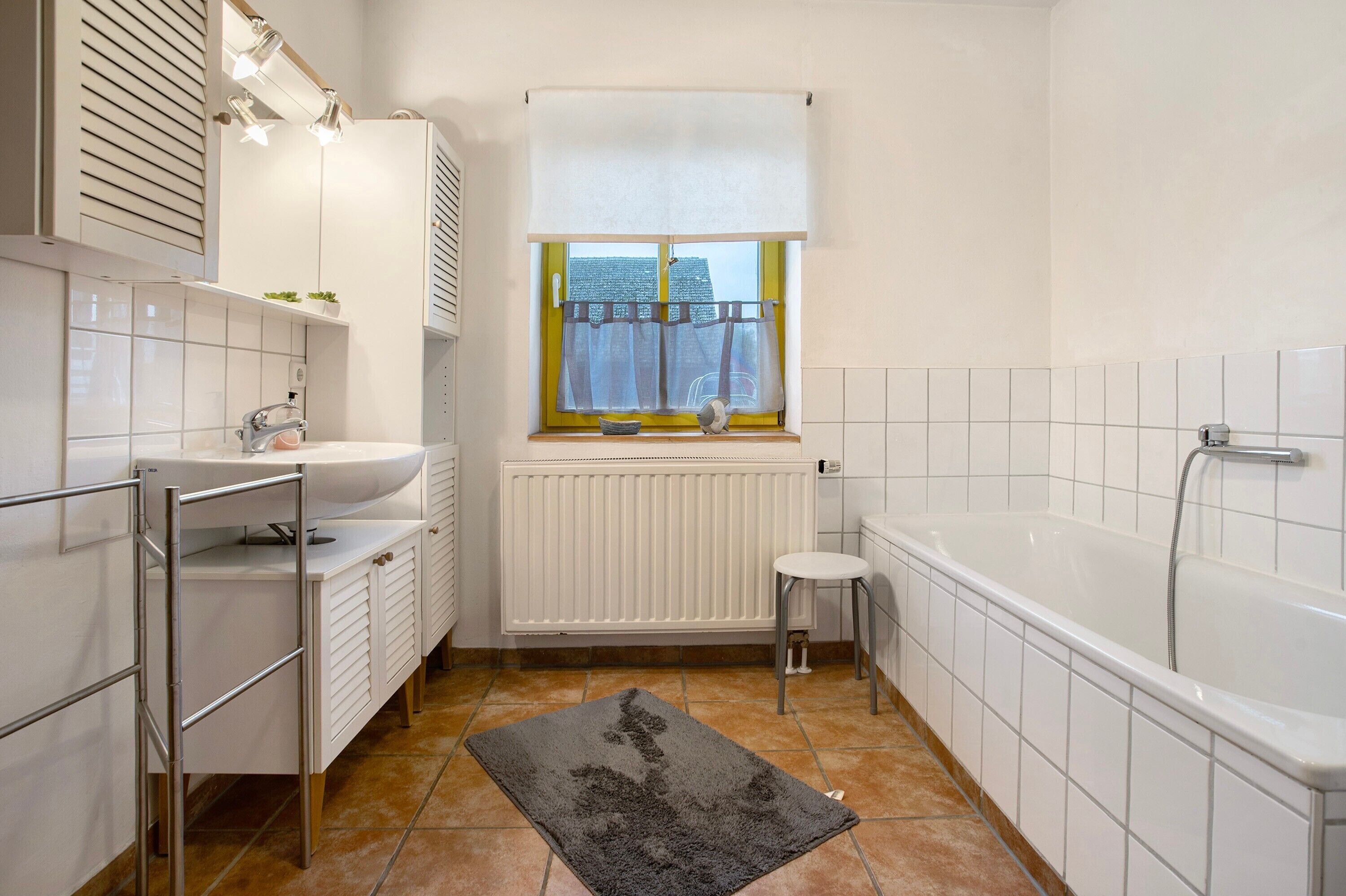 Een badkamer met een bad, een raam met gordijn, een radiator en een wastafel.