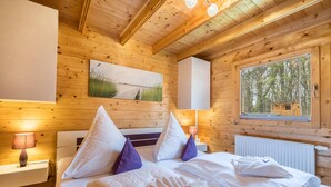 A wooden cabin bedroom with a bed, pillows, a nightstand, a lamp, a painting, and a window with a view of trees. - Holiday Home 'Ferienhäuser Familie Täubert' with Private Terrace, Private Garden and Wi-Fi (Boxberg/O.L.)