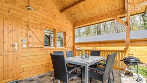 A wooden cabin with a covered patio, a dining table with chairs, a barbecue grill, and a view of the surrounding forest. - Holiday Home 'Ferienhäuser Familie Täubert' with Private Terrace, Private Garden and Wi-Fi (Boxberg/O.L.)