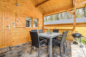 Une cabane en bois dotée d’un patio couvert, d’une table à manger avec des chaises, d’un barbecue et offrant une vue sur la forêt environnante.