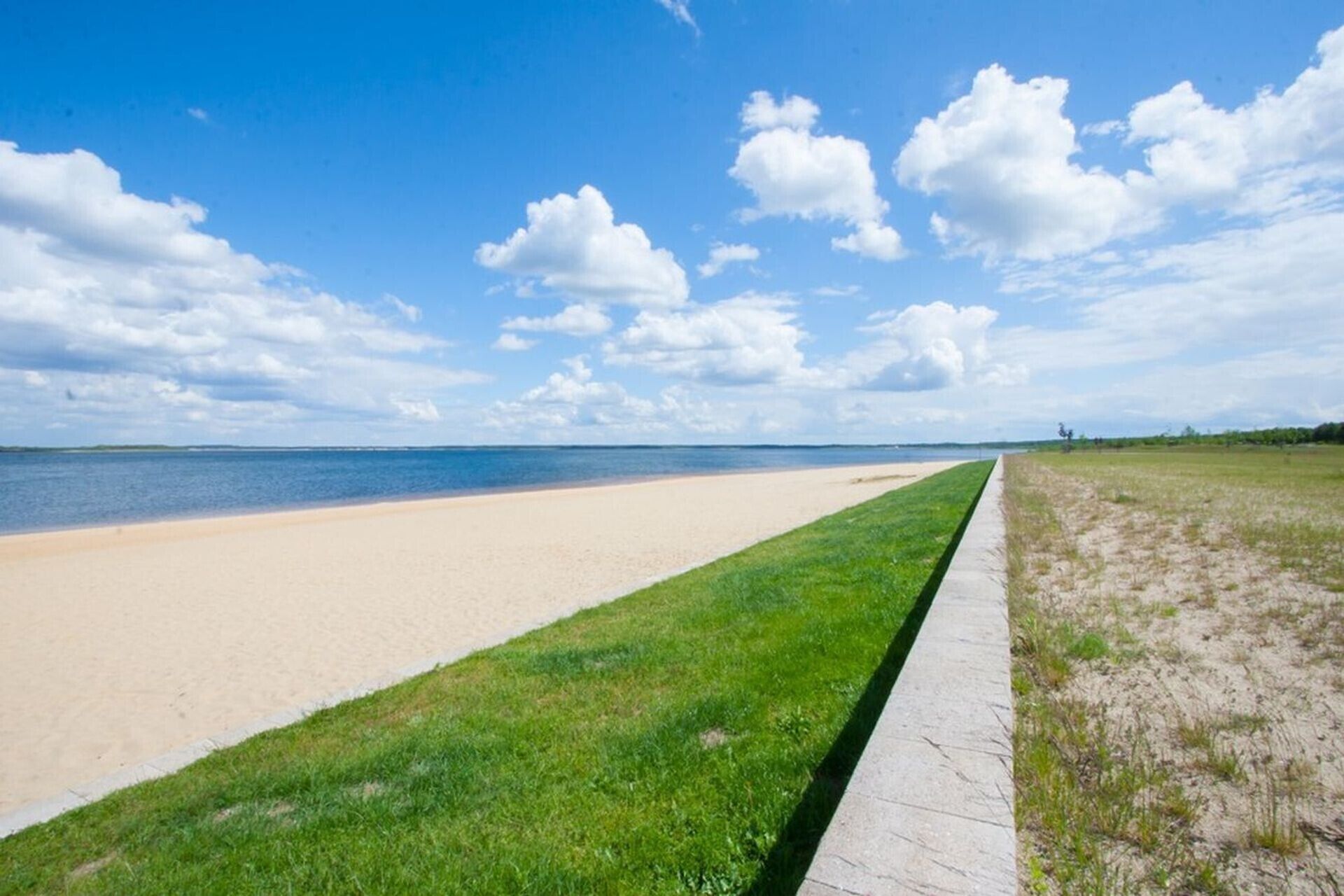 Une plage avec un sentier bordé d’herbe menant à l’eau, un ciel dégagé et une terre lointaine à l’horizon.