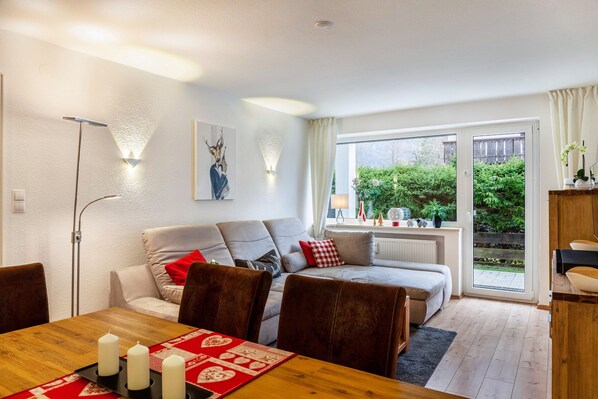 A modern living room with a sofa, dining table, and chairs, a floor lamp, and a view of greenery outside.