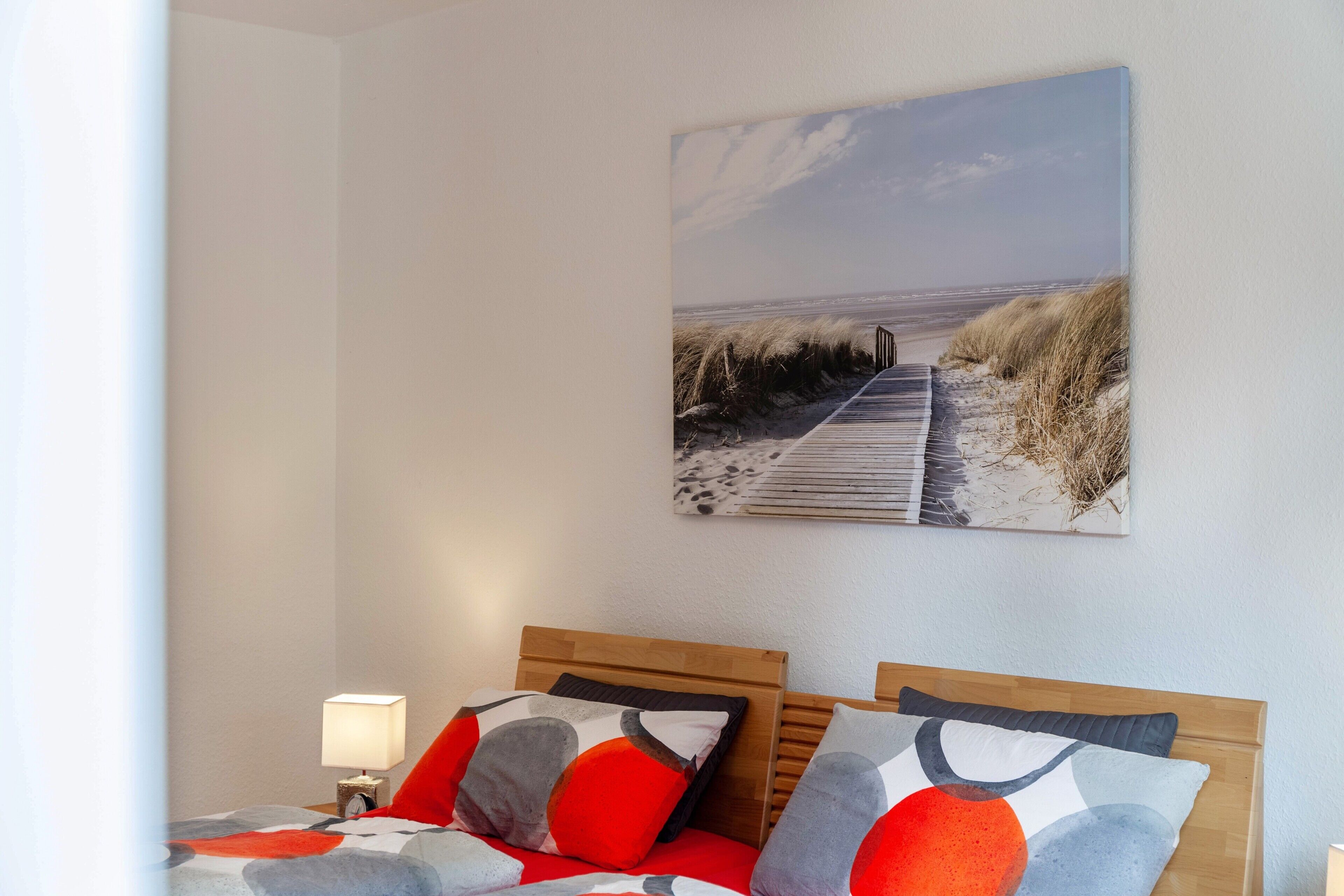 Et hotelværelse med sengegavl i træ, en seng med røde og grå puder, et natbord med en lampe og et maleri af en strandscene på væggen.