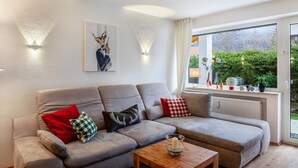 A modern living room with a grey sofa, red and black cushions, a wooden coffee table, and a window with a view of greenery. - Apartment 'Ferienwohnung Alpenglück' with Mountain View, Terrace and Wi-Fi (Mittenwald)